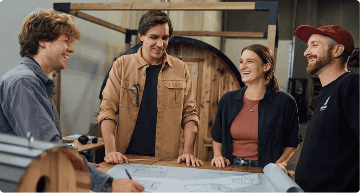 A business owner with his employees gathered around a table looking at design plans while looking at each other and smiling.